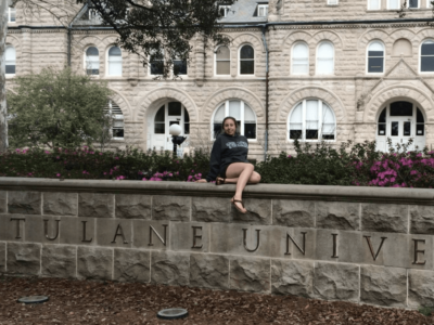 girl in a Tulane hoodie sitting on the sign that says "Tulane University" outside Tulane's Gibson Hall