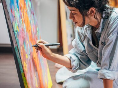 A girl in the arts major sitting on the ground painting on a canvas.