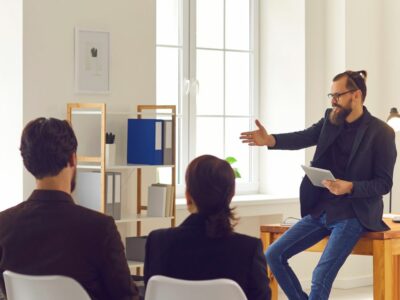 Someone in management leading a discussion in front of peers sitting in white chairs